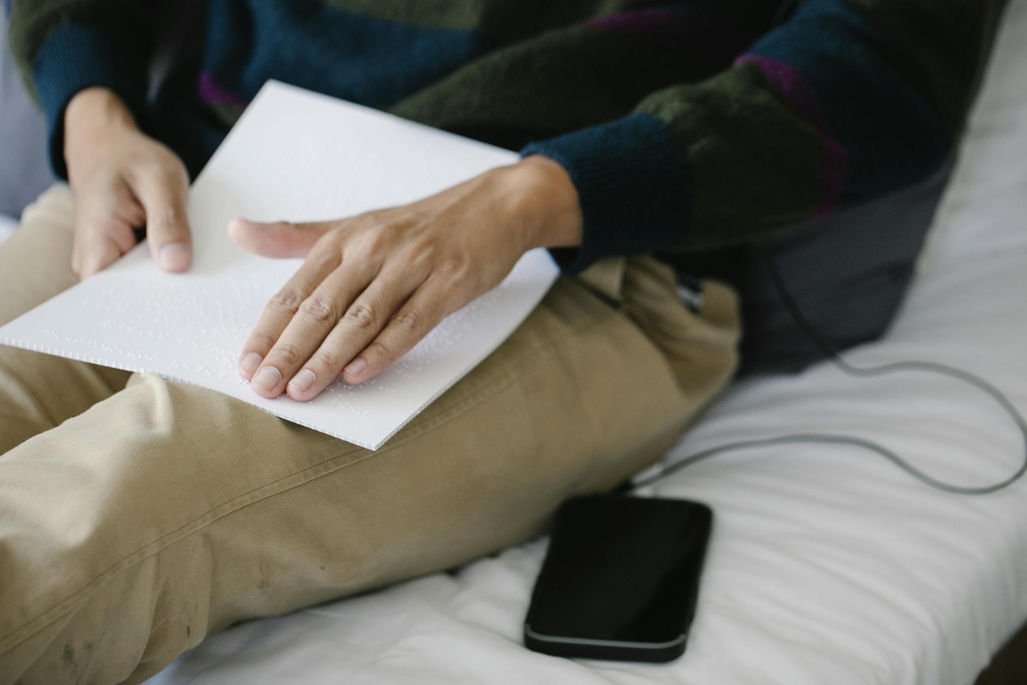 A person reads Braille text while sitting on a bed with a smartphone nearby.