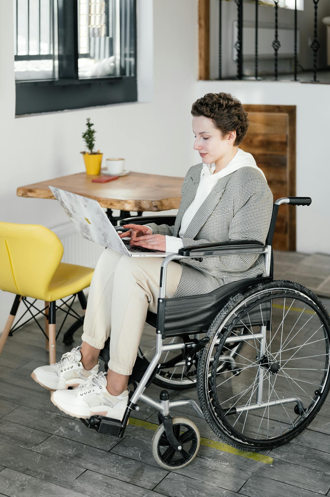 A young woman in a wheelchair working on a laptop in a modern cafe setting.