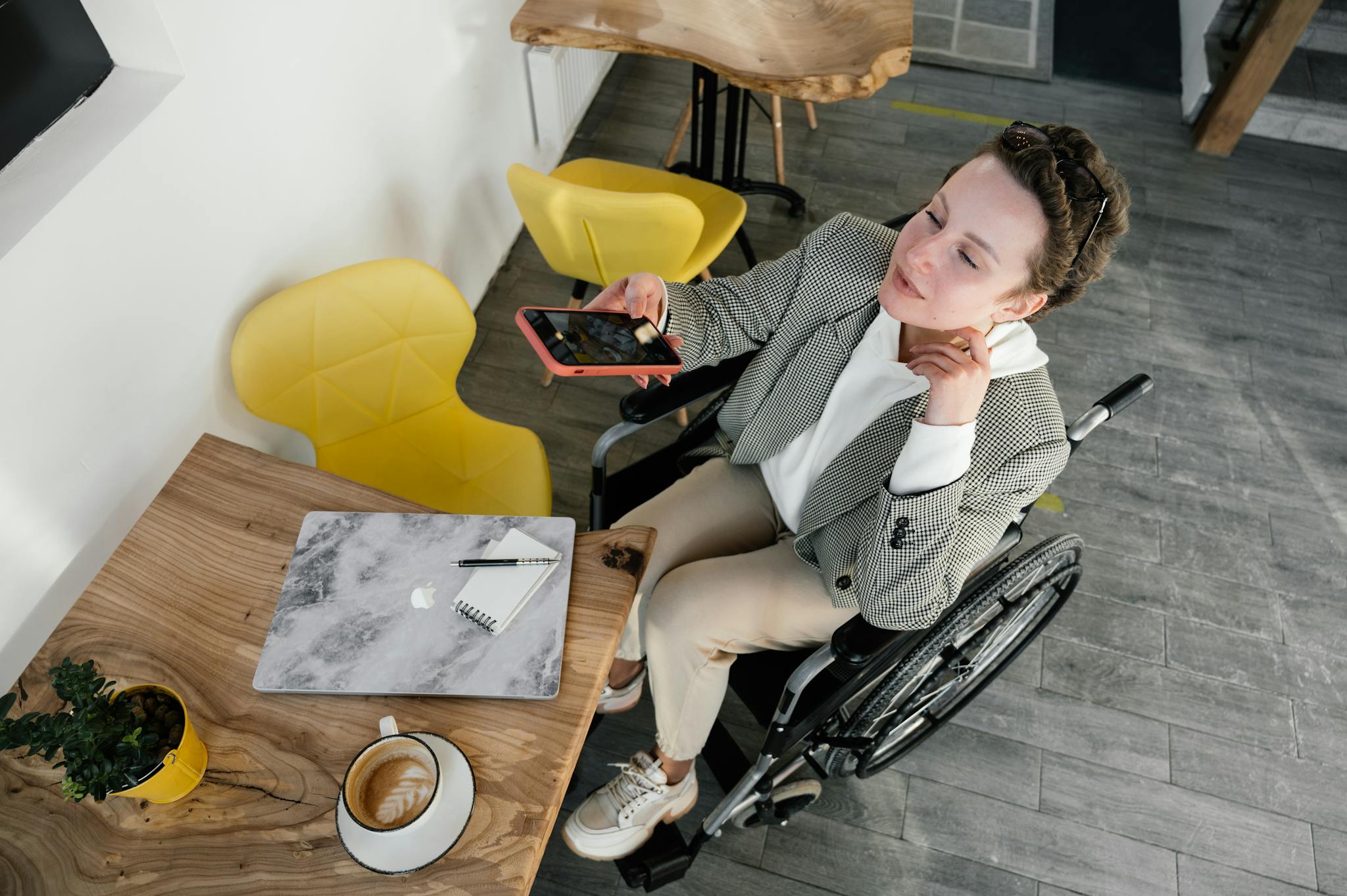 High-angle view of a woman in a wheelchair working on a laptop and phone in a café. Inclusive workspace concept.
